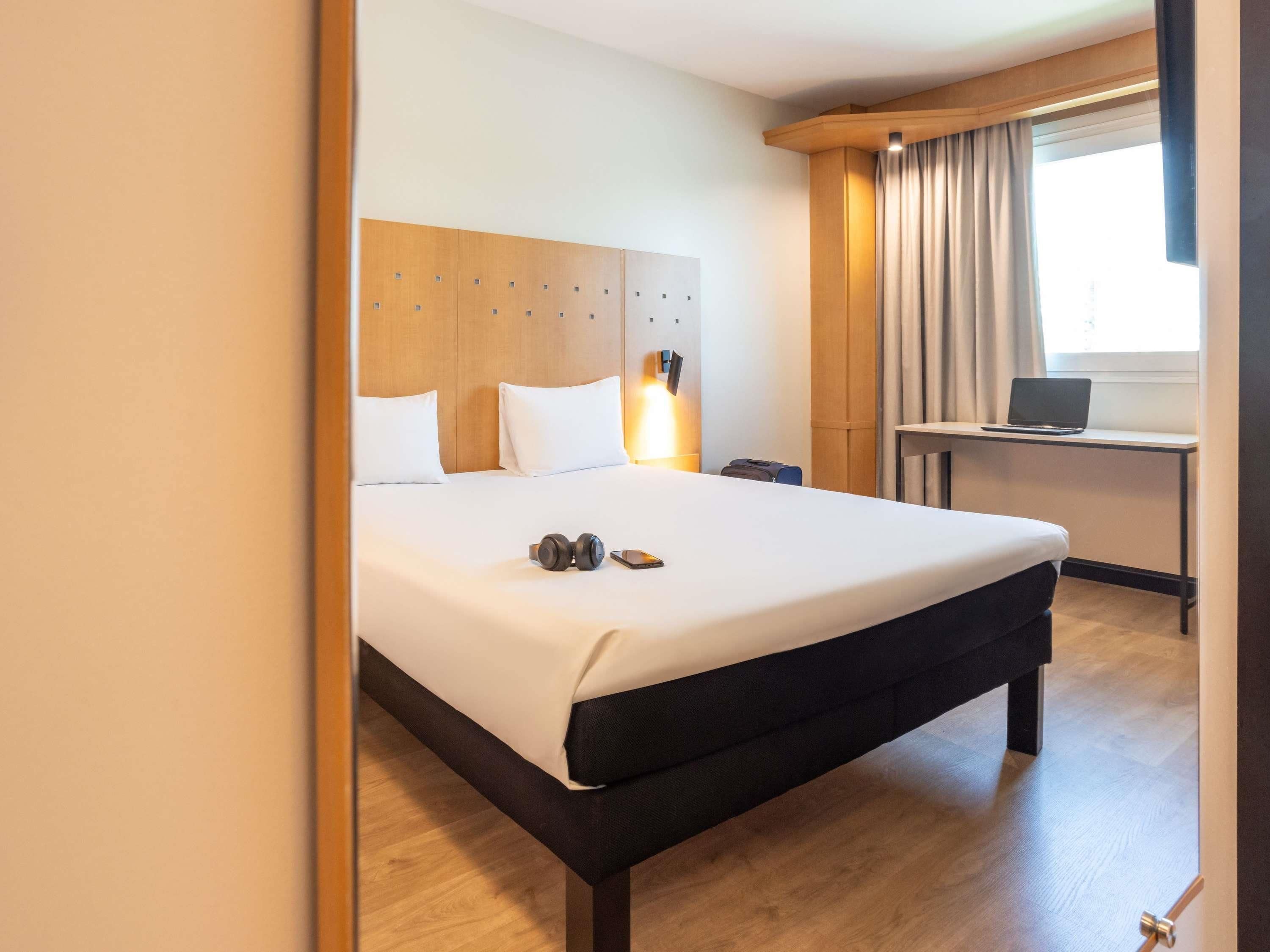 Una habitación de hotel con una cama grande, un escritorio con una computadora portátil y una ventana con cortinas.