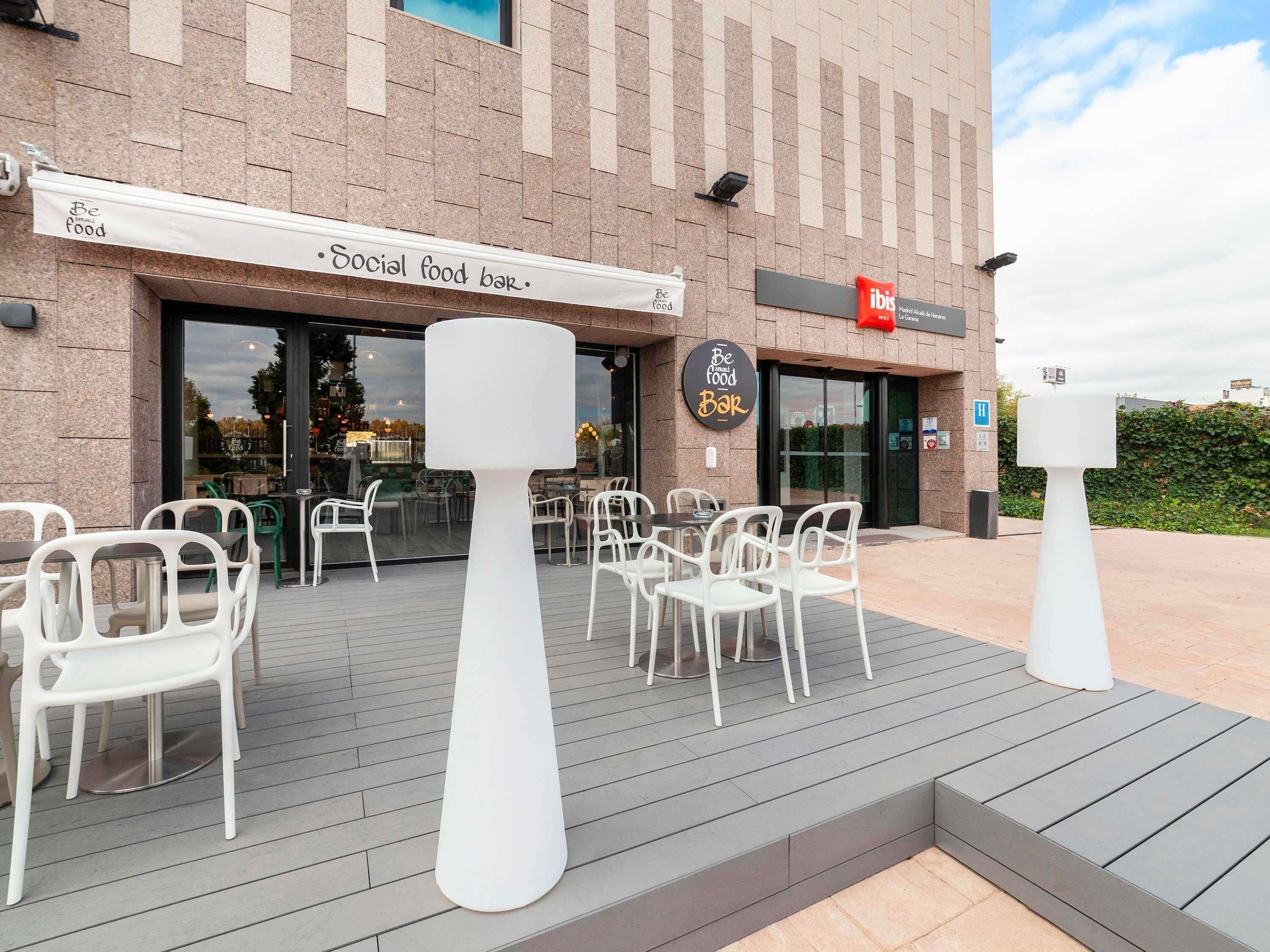Un espace extérieur aménagé avec des chaises et des tables blanches, situé devant un bâtiment sur lequel est apposée une enseigne indiquant « Bar proposant une cuisine conviviale ».