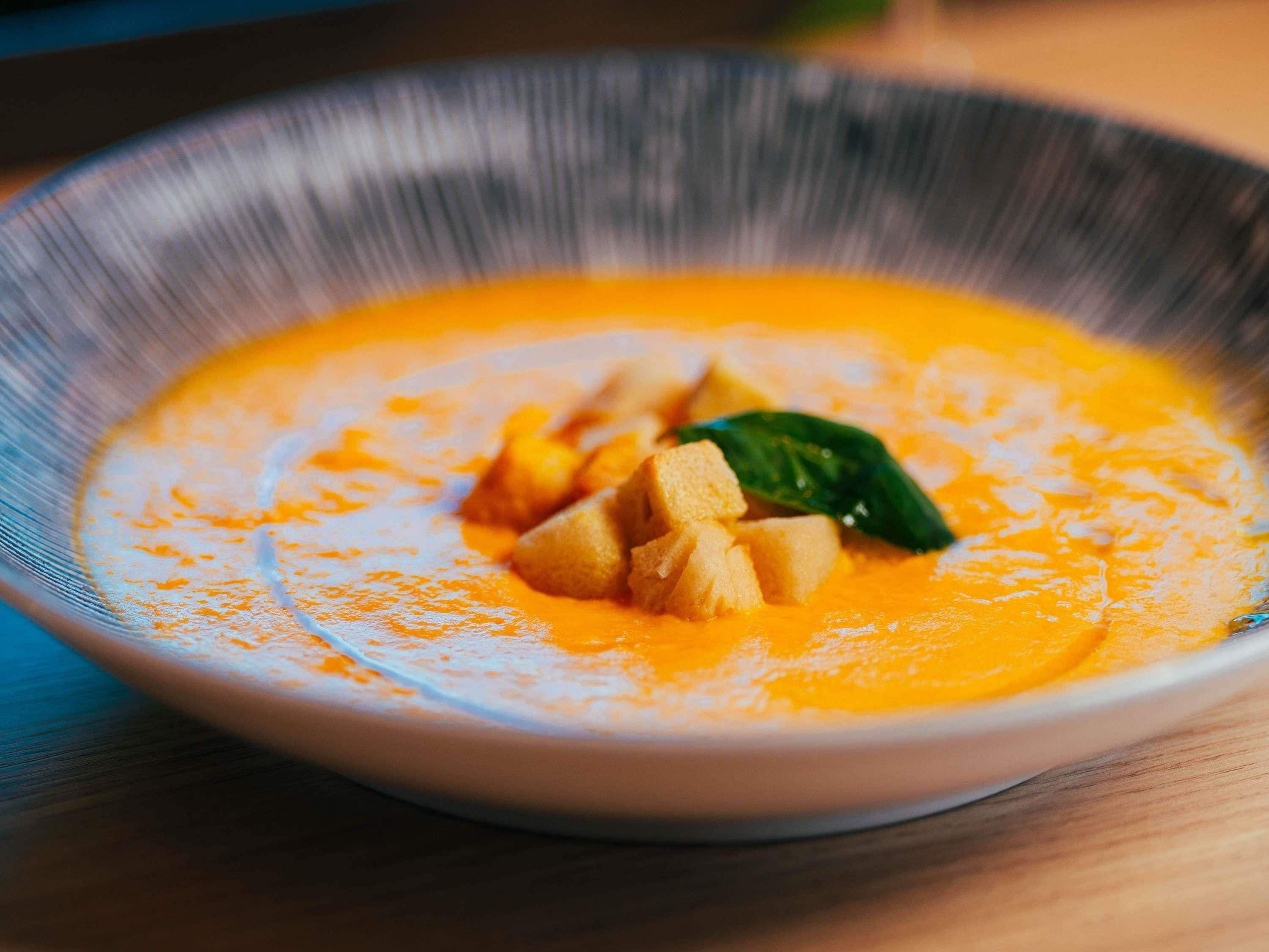 Un tazón de sopa de color naranja cremoso, adornada con una hoja verde y cubos de comida encima.