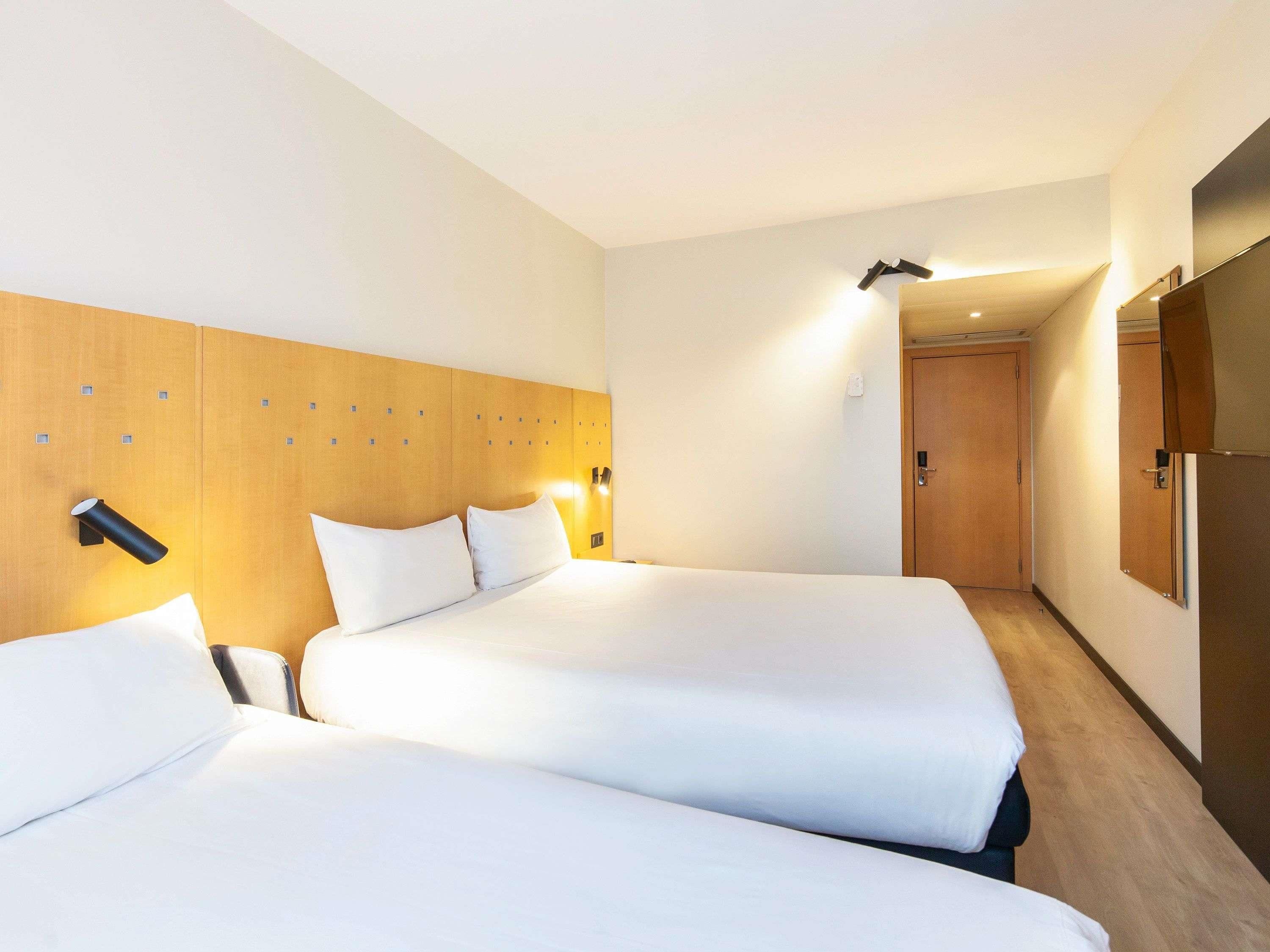 Een hotelkamer met twee bedden, een houten hoofdeinde, nachtlampjes en een deur naar een andere kamer.