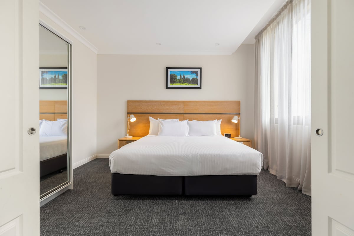 Een hotelkamer met een groot bed, een houten hoofdbord, twee bedlampen, een lijst met een afbeelding aan de muur, en uitzicht op de stad door het raam.
