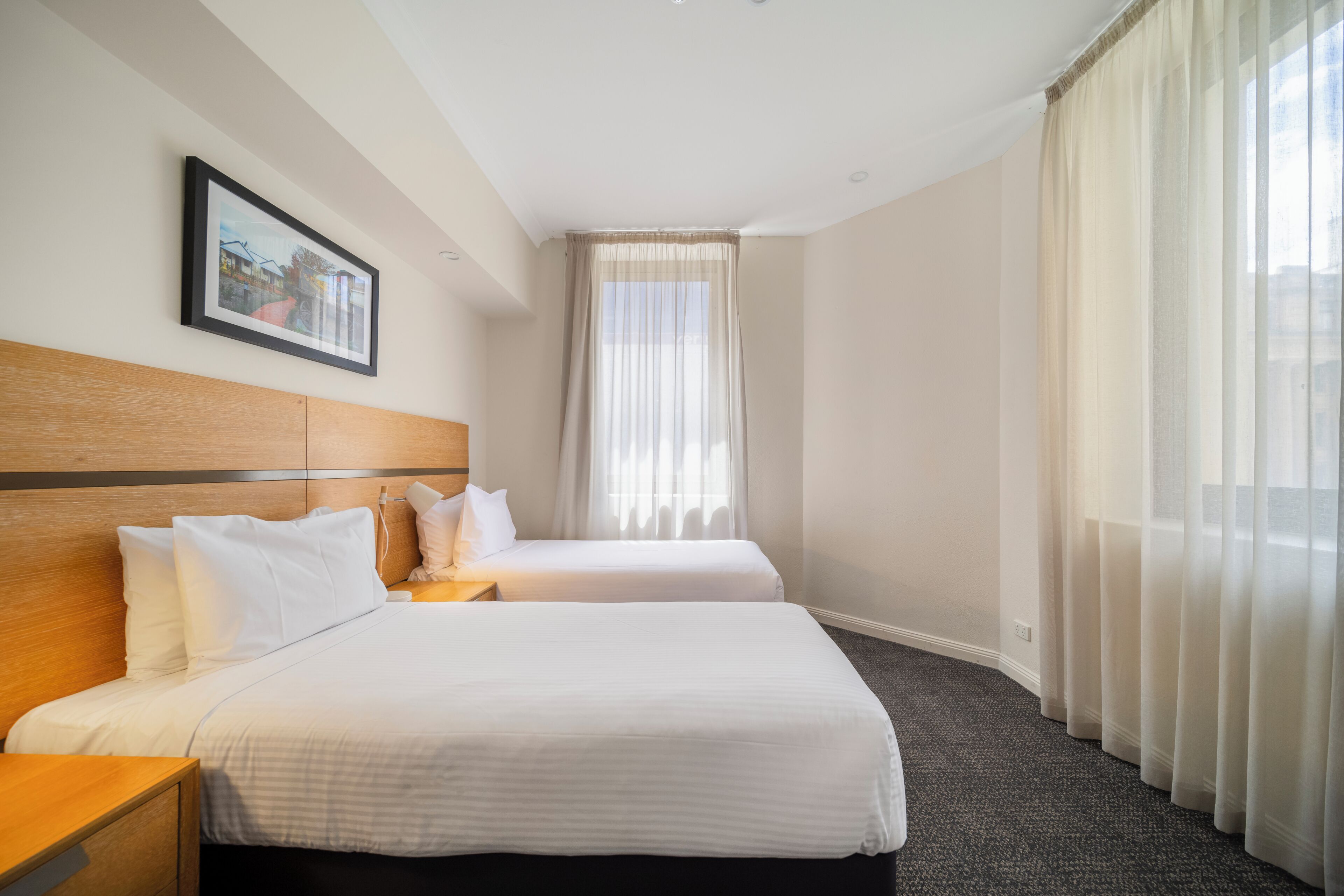 A hotel room with two beds, a wooden headboard, a nightstand, a framed picture on the wall, and sheer curtains.