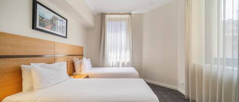 A hotel room with two beds, a wooden headboard, a nightstand, a framed picture on the wall, and sheer curtains.