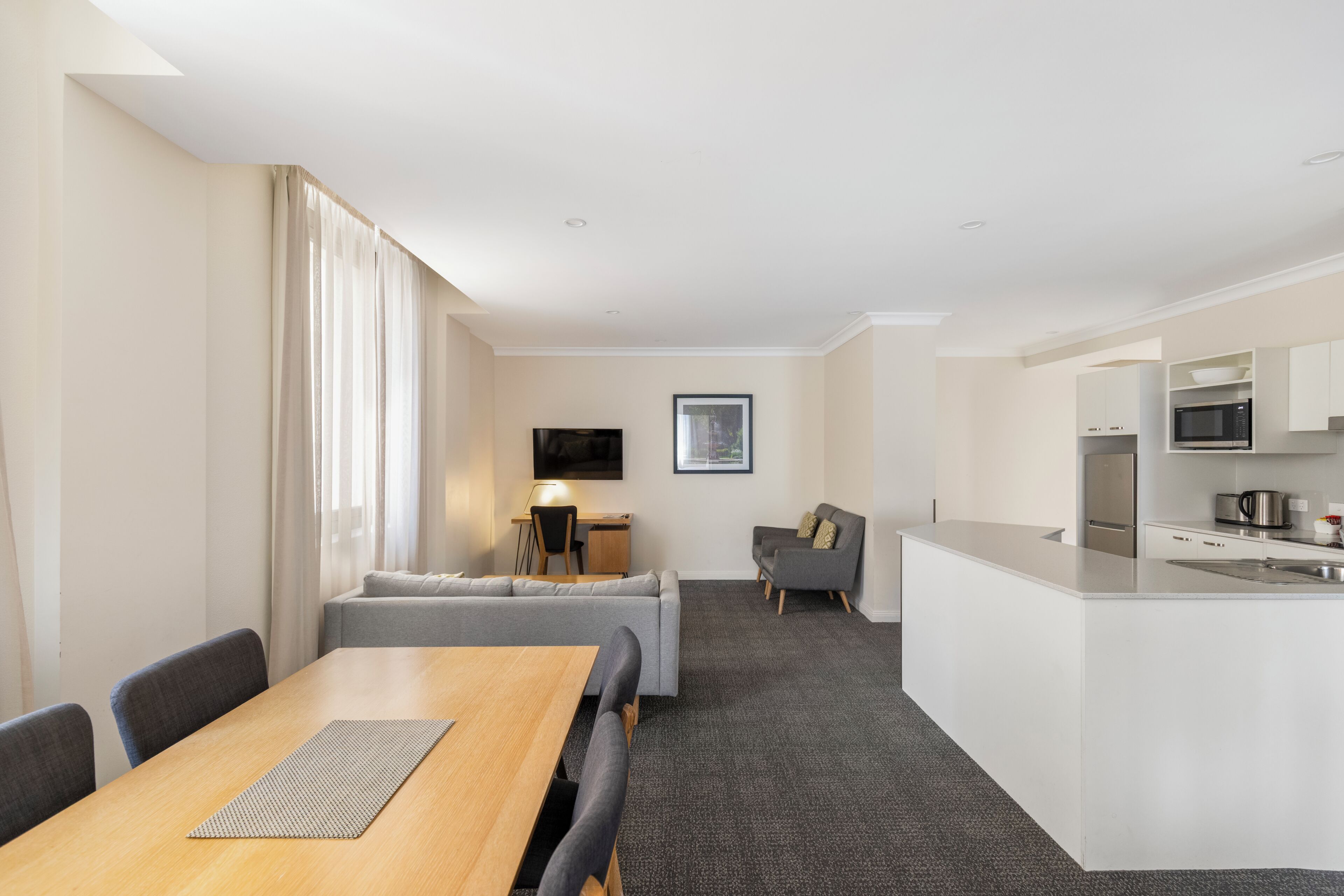 A modern hotel room with a dining table, sofa, kitchenette, and a flat-screen TV.