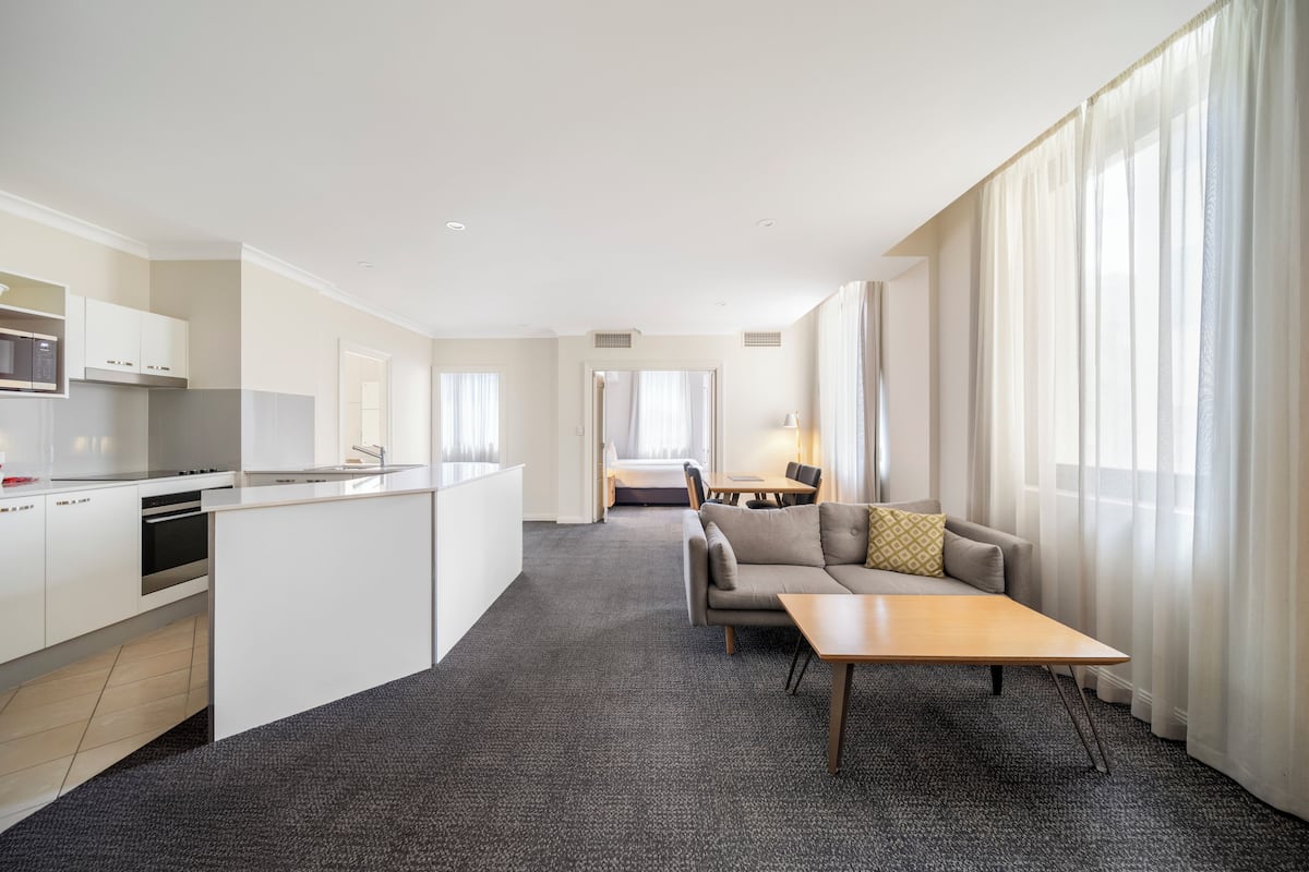 Een moderne hotelkamer met een kitchenette, een bank, een eettafel en grote ramen met lichte gordijnen.
