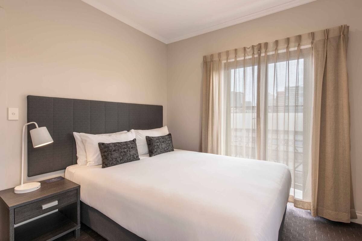 A hotel room with a large bed, a bedside table with a lamp, and a view through sheer curtains.