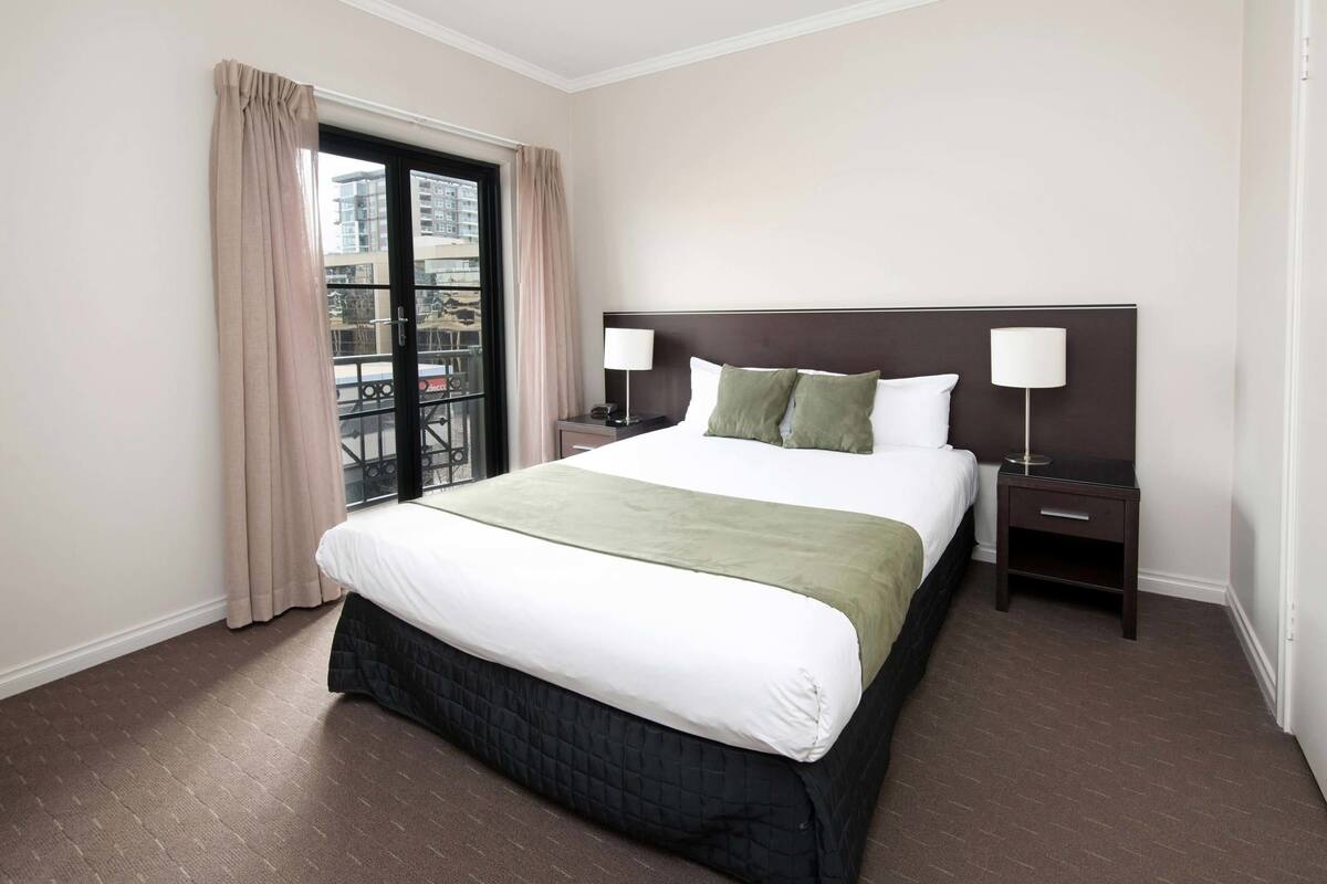 A hotel room with a large bed, two bedside tables, a window with curtains, and a view of buildings outside.