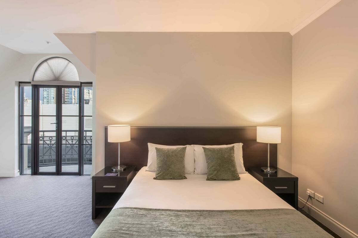 A hotel room with a large bed, two bedside lamps, and a view of the outside through a glass door.