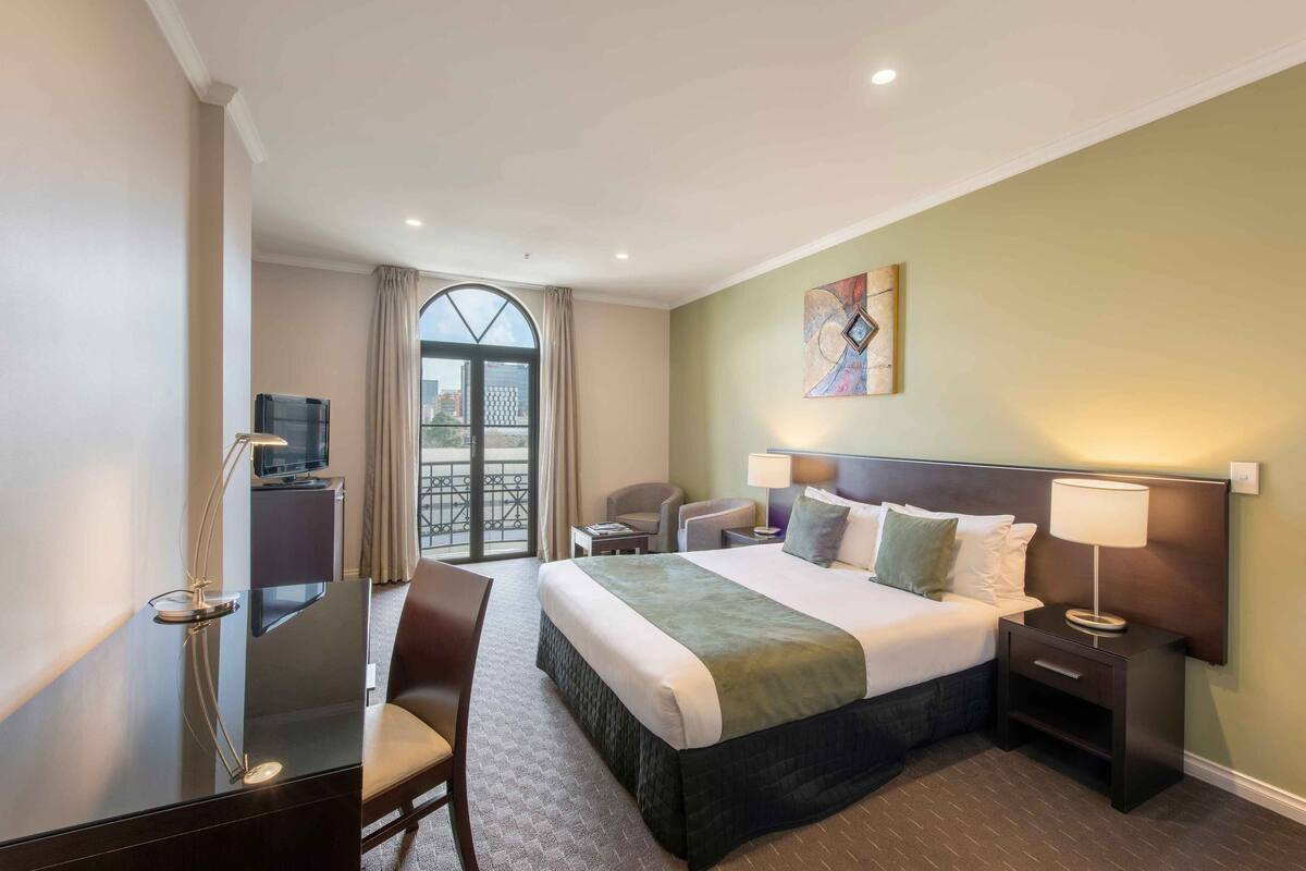 A hotel room with a bed, bedside tables, a desk, a chair, a TV, and a window with curtains.