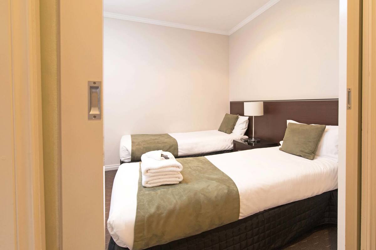 A hotel room with two beds, each with a green and white bedspread, a folded white towel on the left bed, and a bedside lamp on the right.