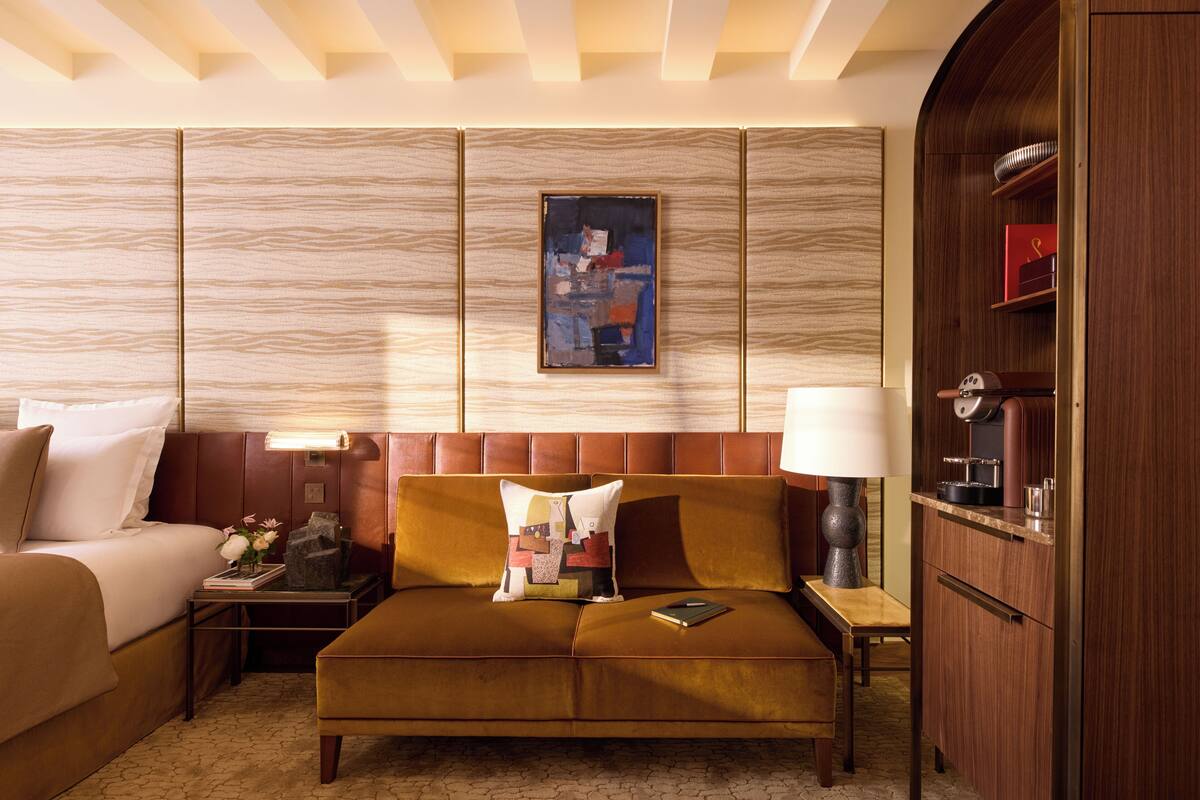 A hotel room with a sofa, a bedside table with a lamp, a framed artwork on the wall, and a wooden cabinet.