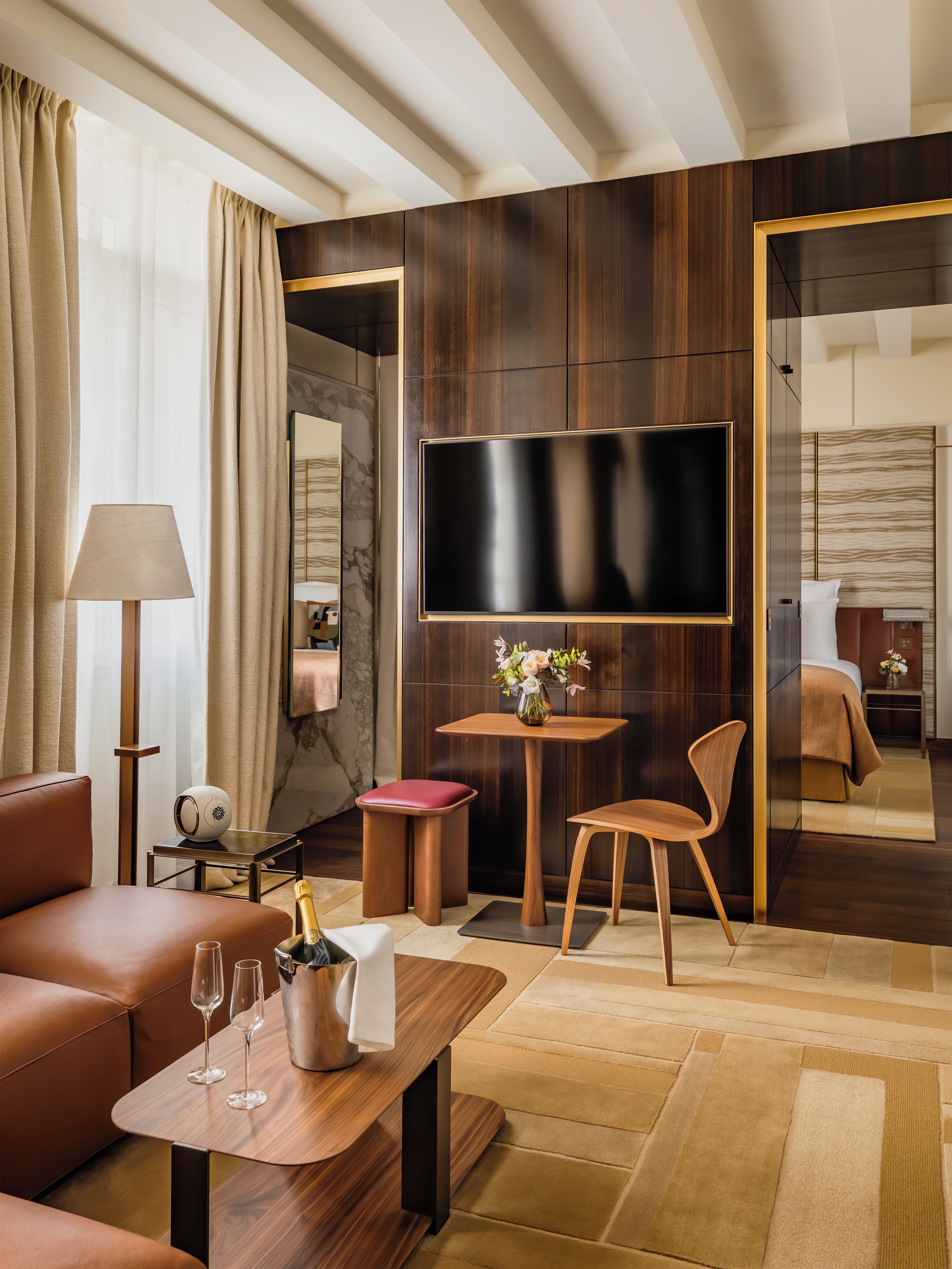 A modern hotel room with a flat-screen TV, leather sofa, wooden coffee table, and a small side table with a lamp and a vase of flowers.