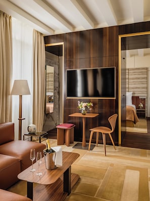 Habitación de hotel moderna con televisión de pantalla plana, sofá de cuero, mesa de centro de madera y una mesita auxiliar con lámpara y un jarrón de flores.