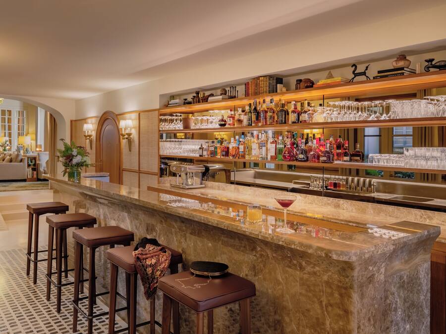 A bar area with a marble countertop, multiple stools, and a variety of bottles and glasses.