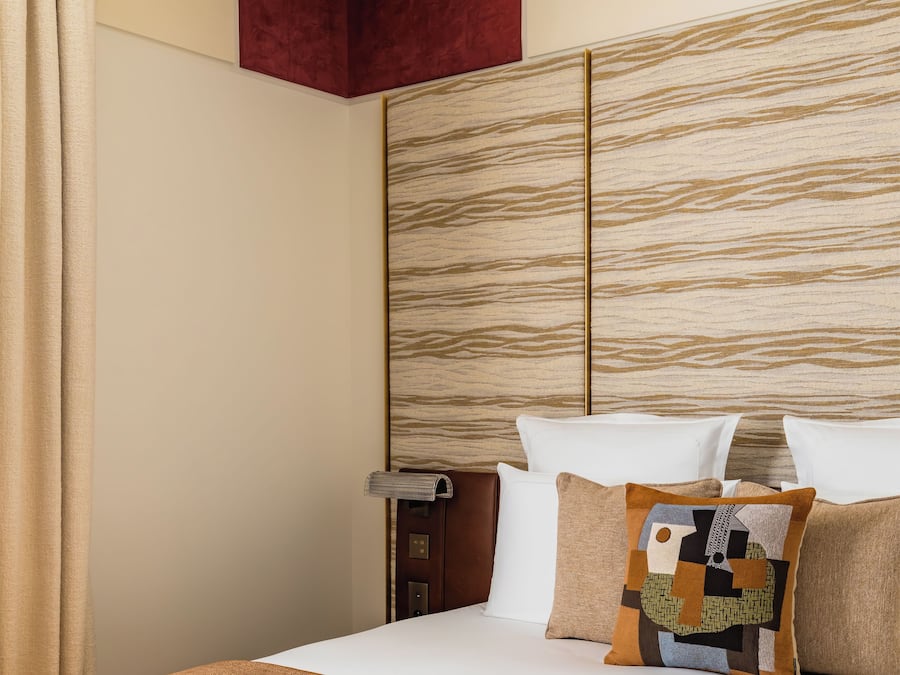 A hotel room with a bed, a wooden headboard, a red accent wall, and a window with curtains.