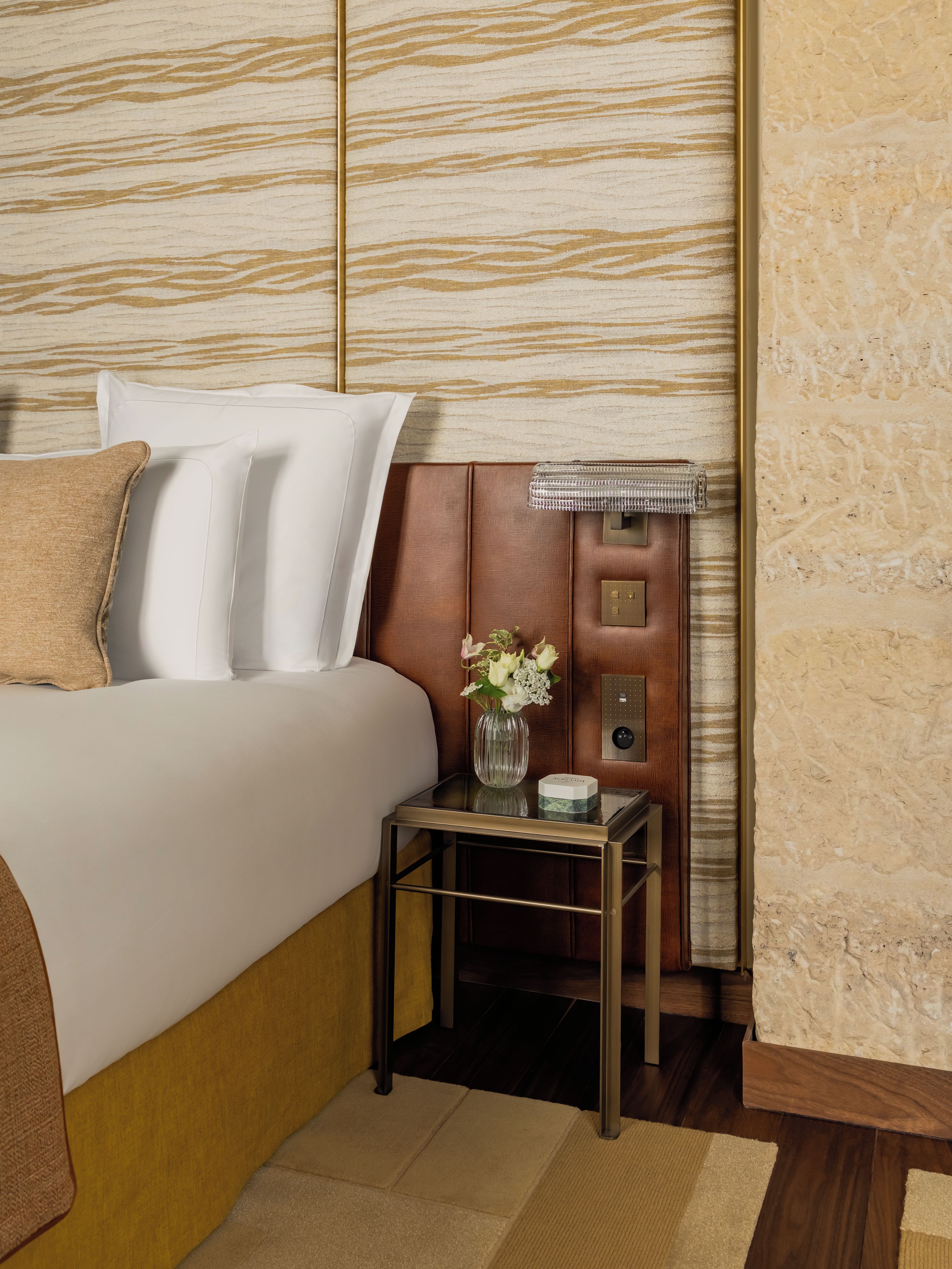 Habitación de hotel con cama, almohadas, mesita de noche con un florero, lámpara y un panel en la pared.