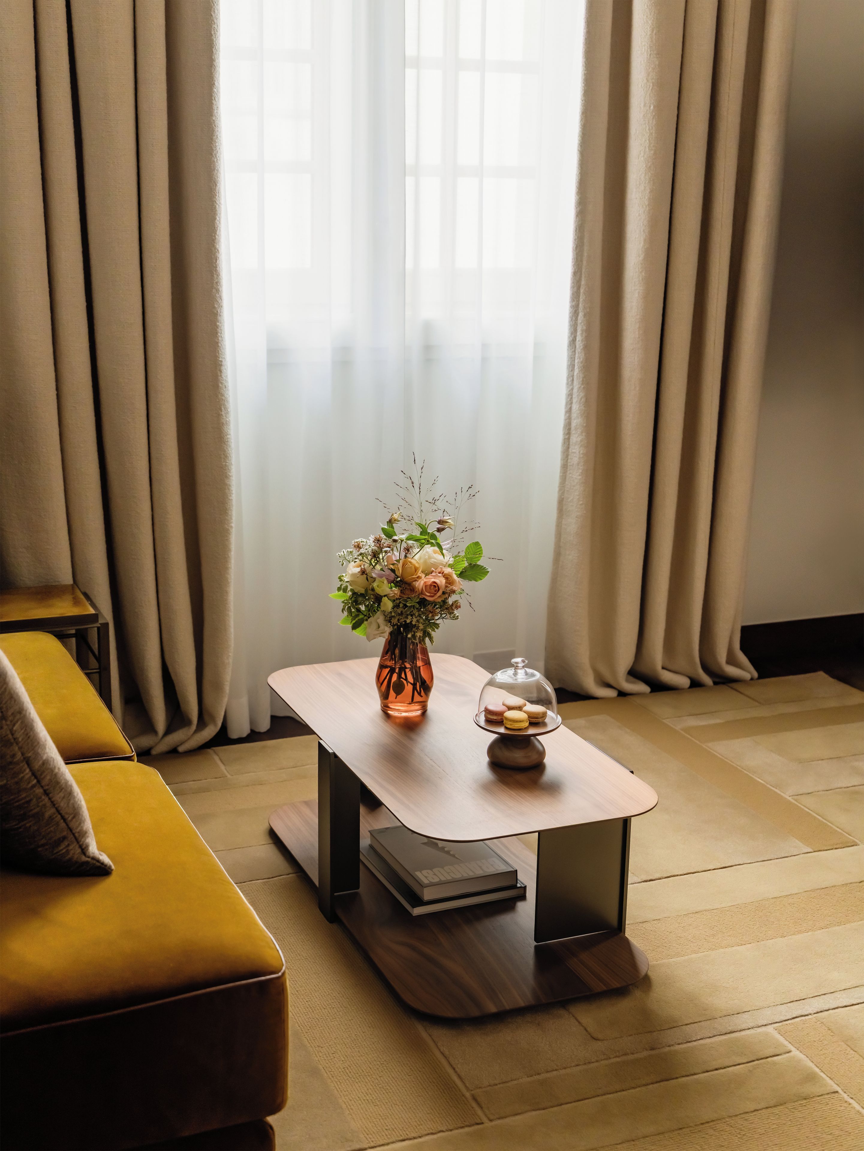 A room with a wooden table, a vase with flowers, and a glass dome with macarons.
