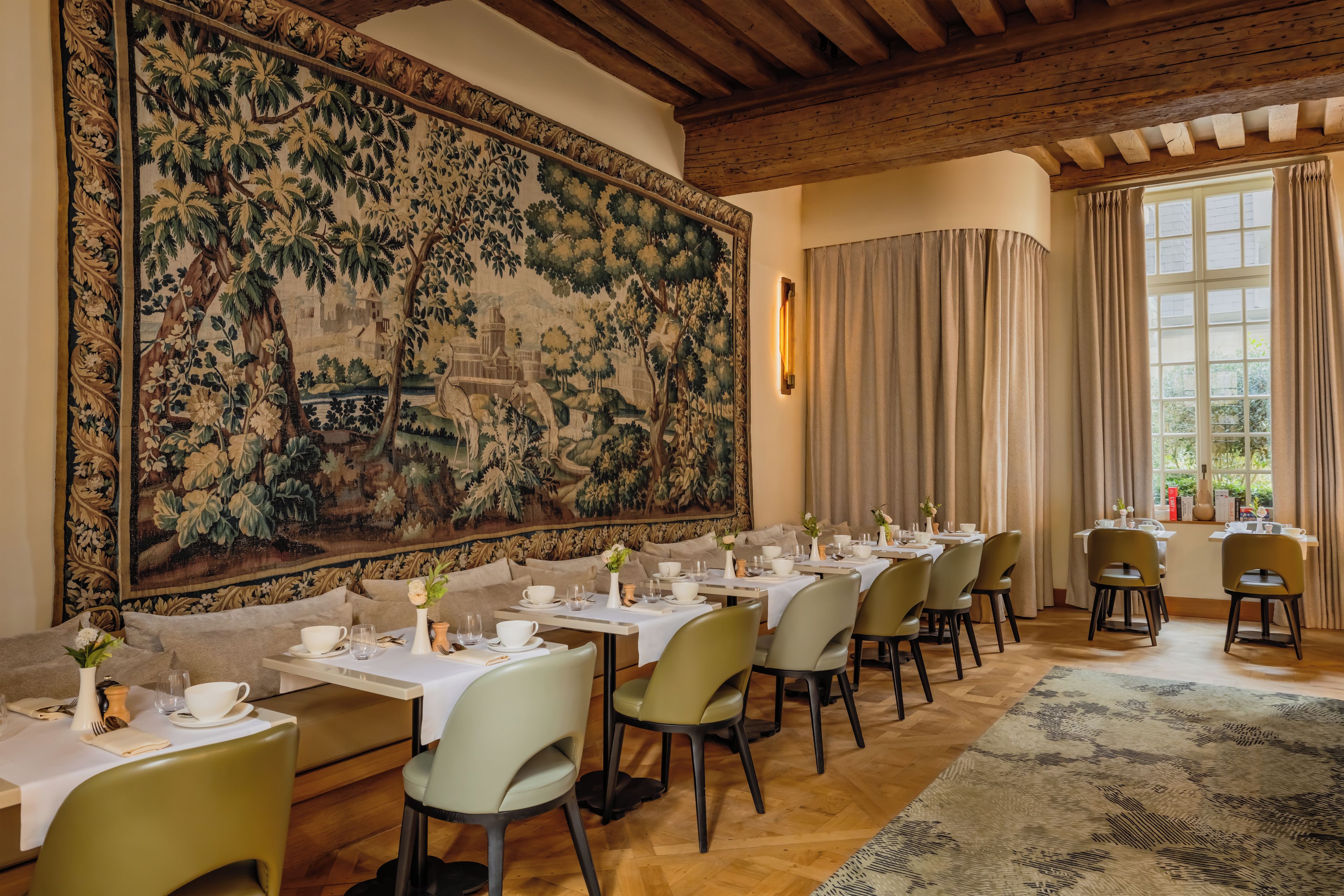 A dining area with tables set for service, a large tapestry on the wall, and a view of the outdoors through a window.