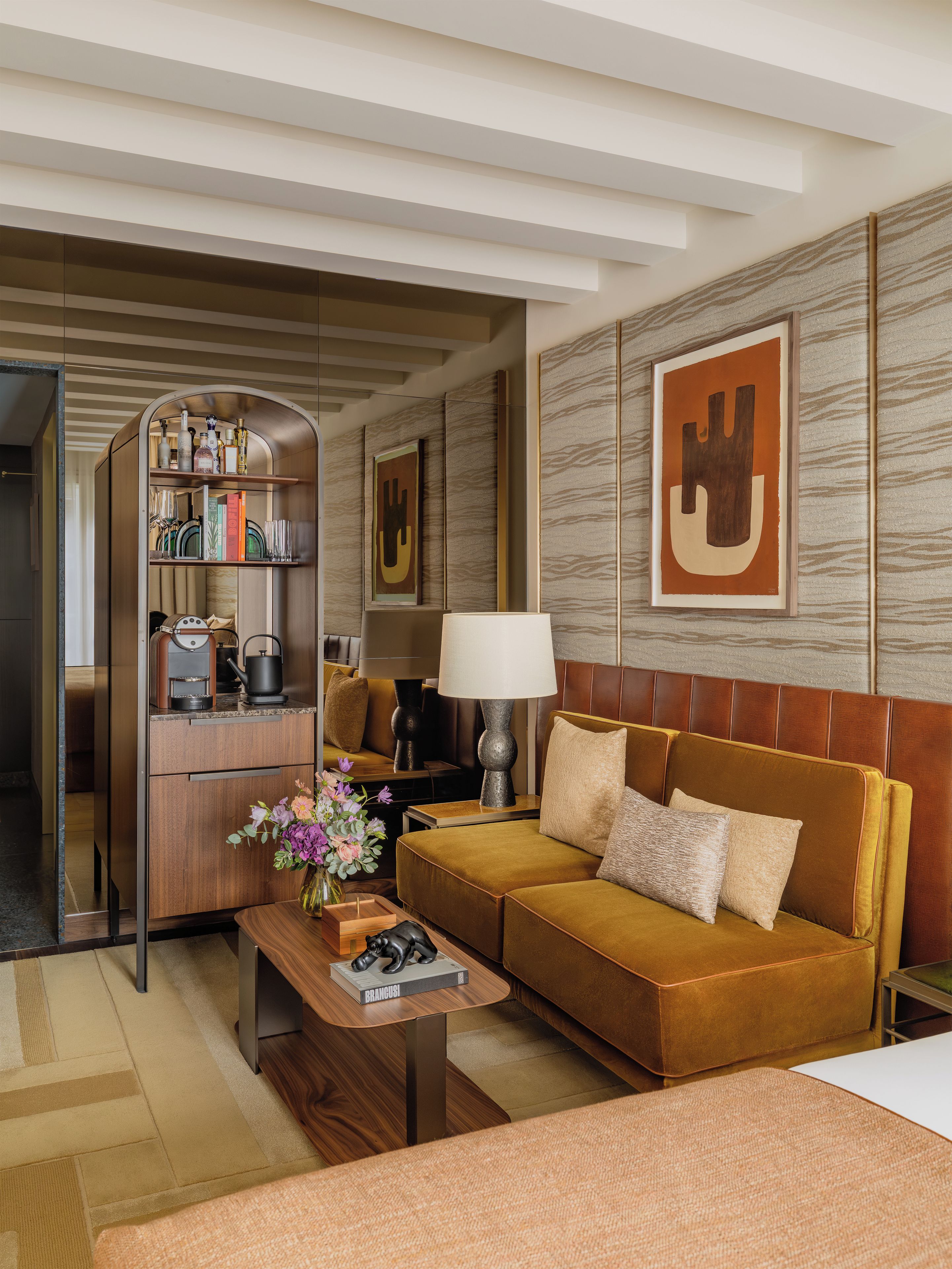 A modern hotel room with a sofa, a coffee table, a bookshelf, and a large abstract painting on the wall.