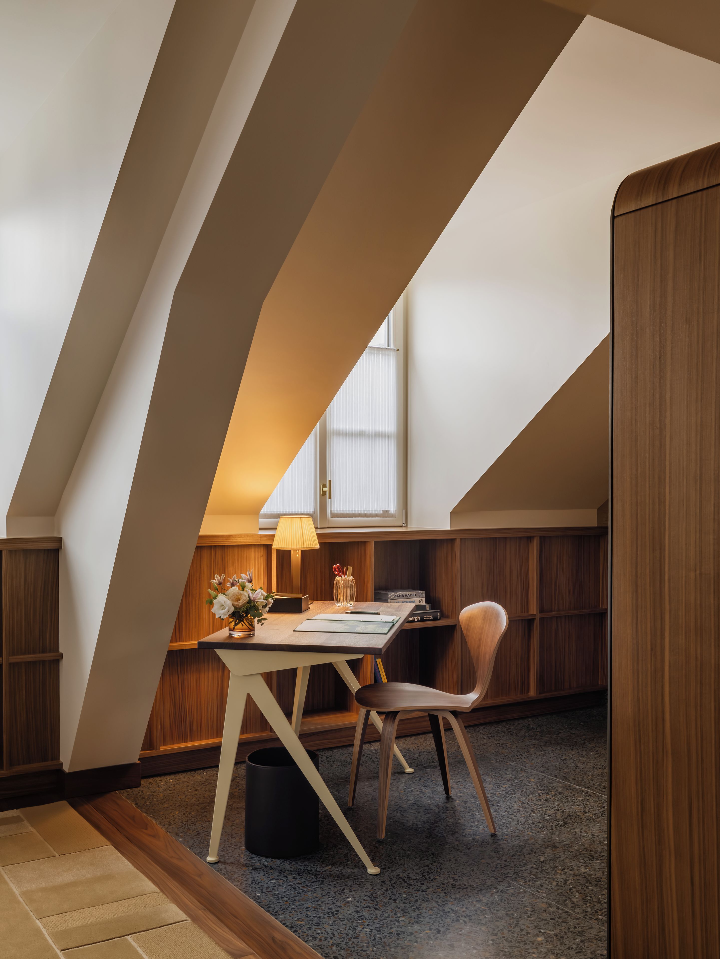 A modern loft bedroom with a sloped ceiling, a wooden desk, a chair, a lamp, and a bookshelf.