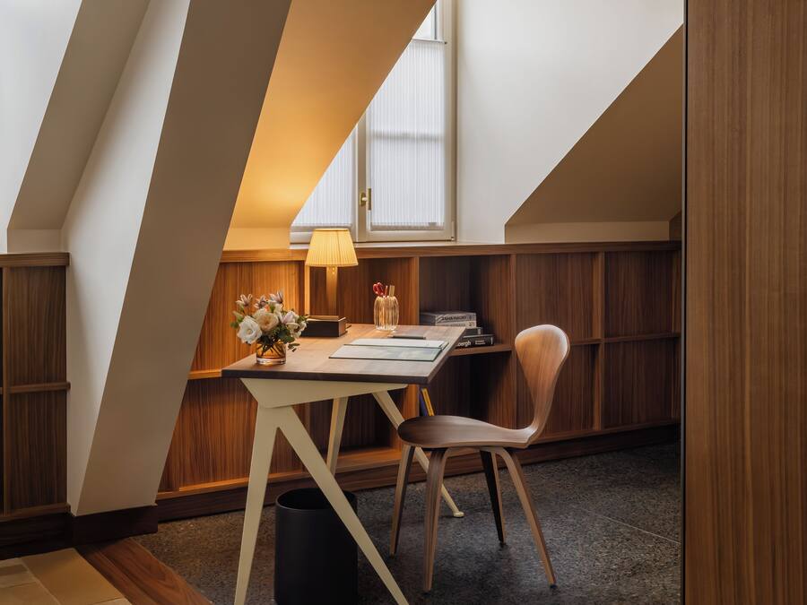 A modern loft bedroom with a sloped ceiling, a wooden desk, a chair, a lamp, and a bookshelf.