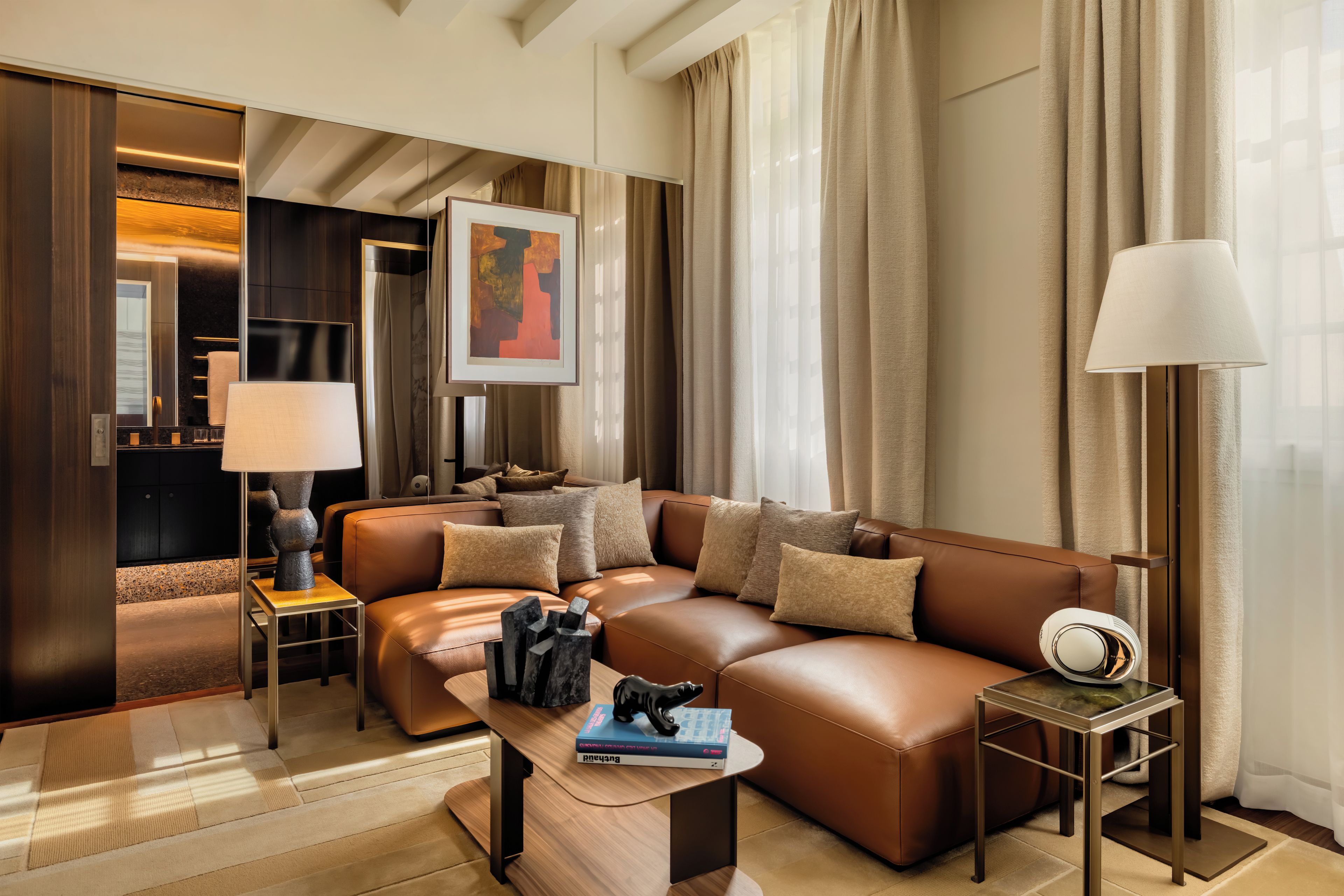 A modern living room with a brown leather sofa, a coffee table with books, a side table with a lamp, and a painting on the wall.