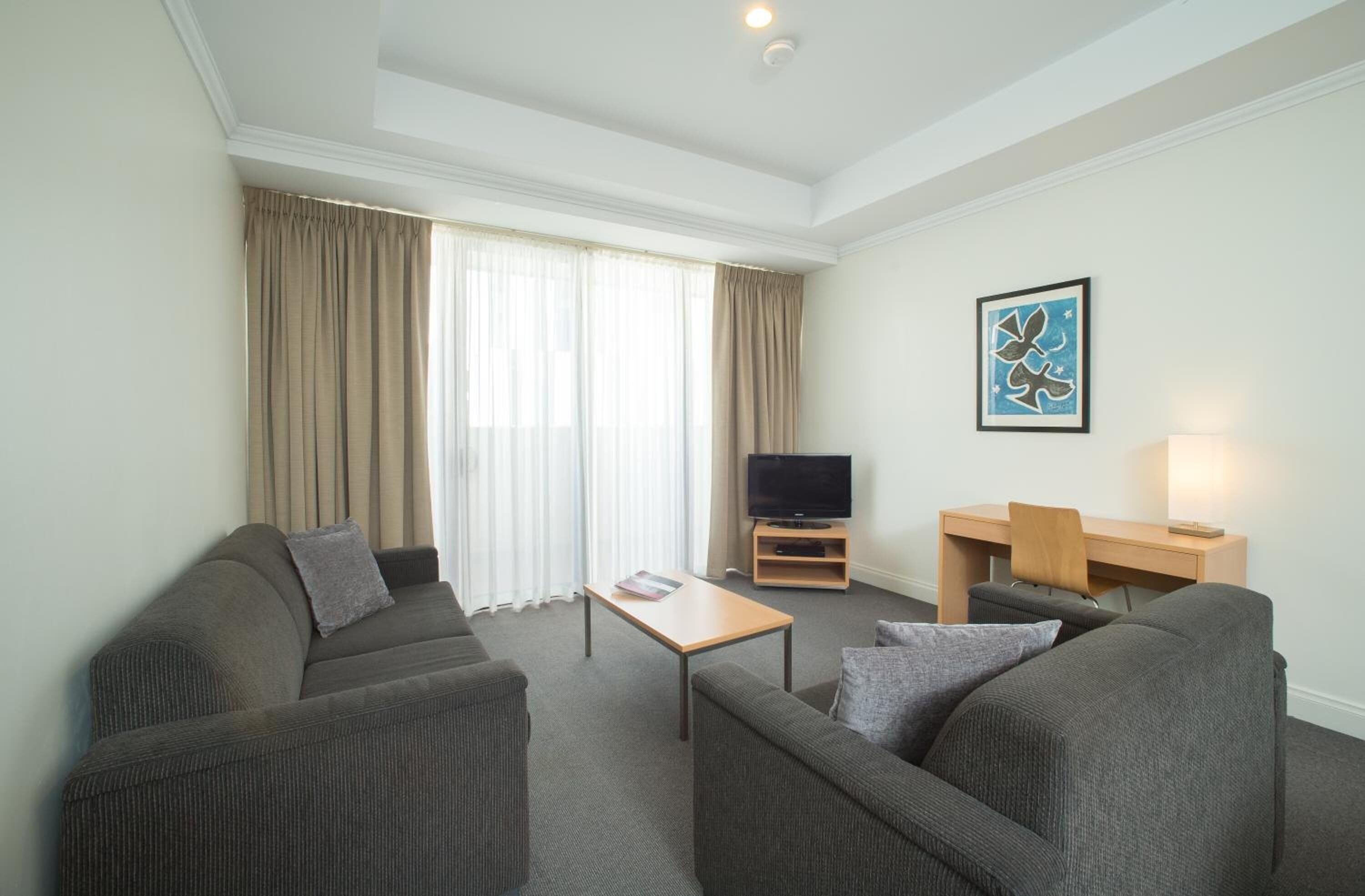 Une chambre d’hôtel avec un canapé gris, une table basse en bois, un téléviseur posé sur un meuble, un bureau avec une chaise et un tableau accroché au mur.
