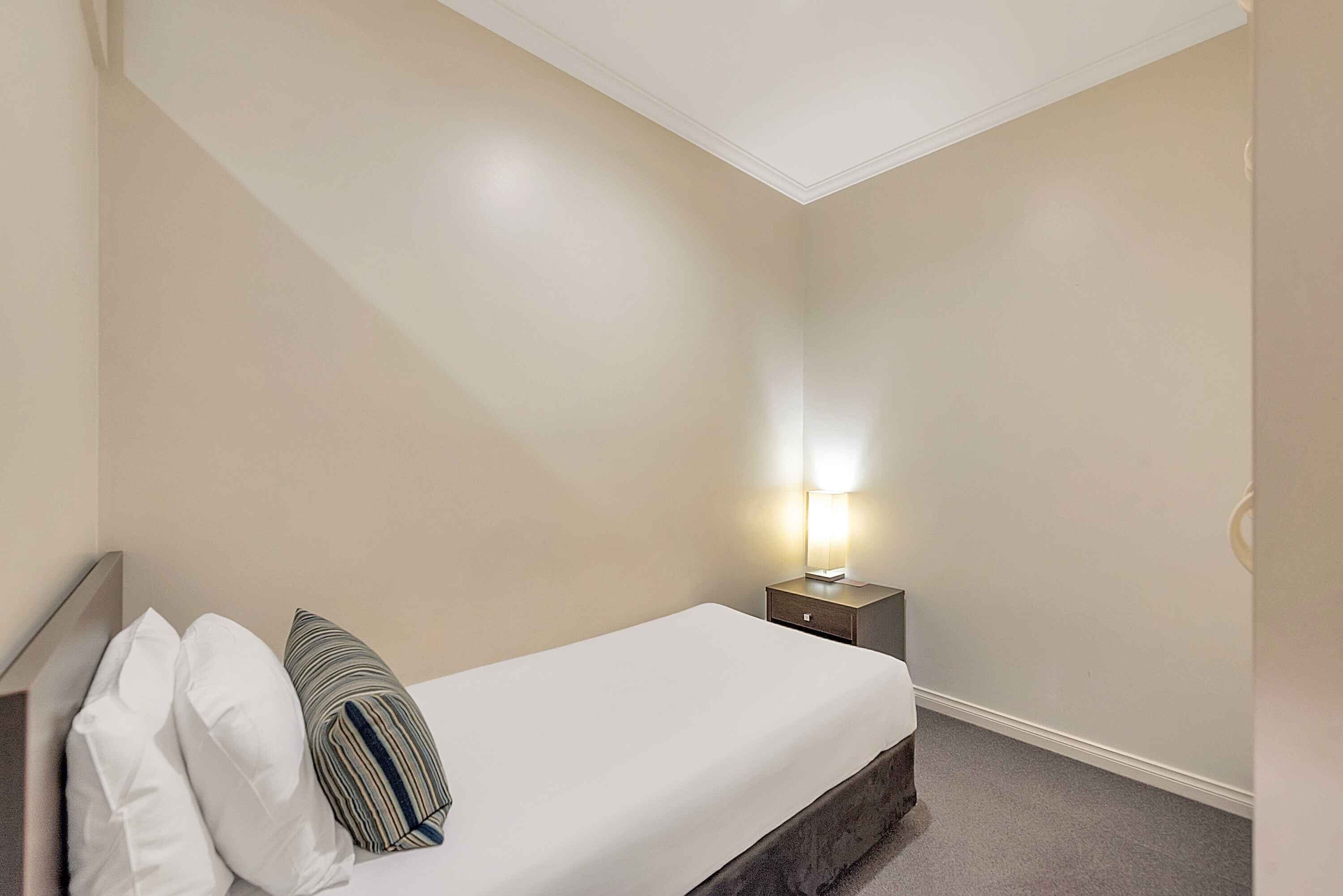 A hotel room with a single bed, a nightstand with a lamp, and a plain wall.
