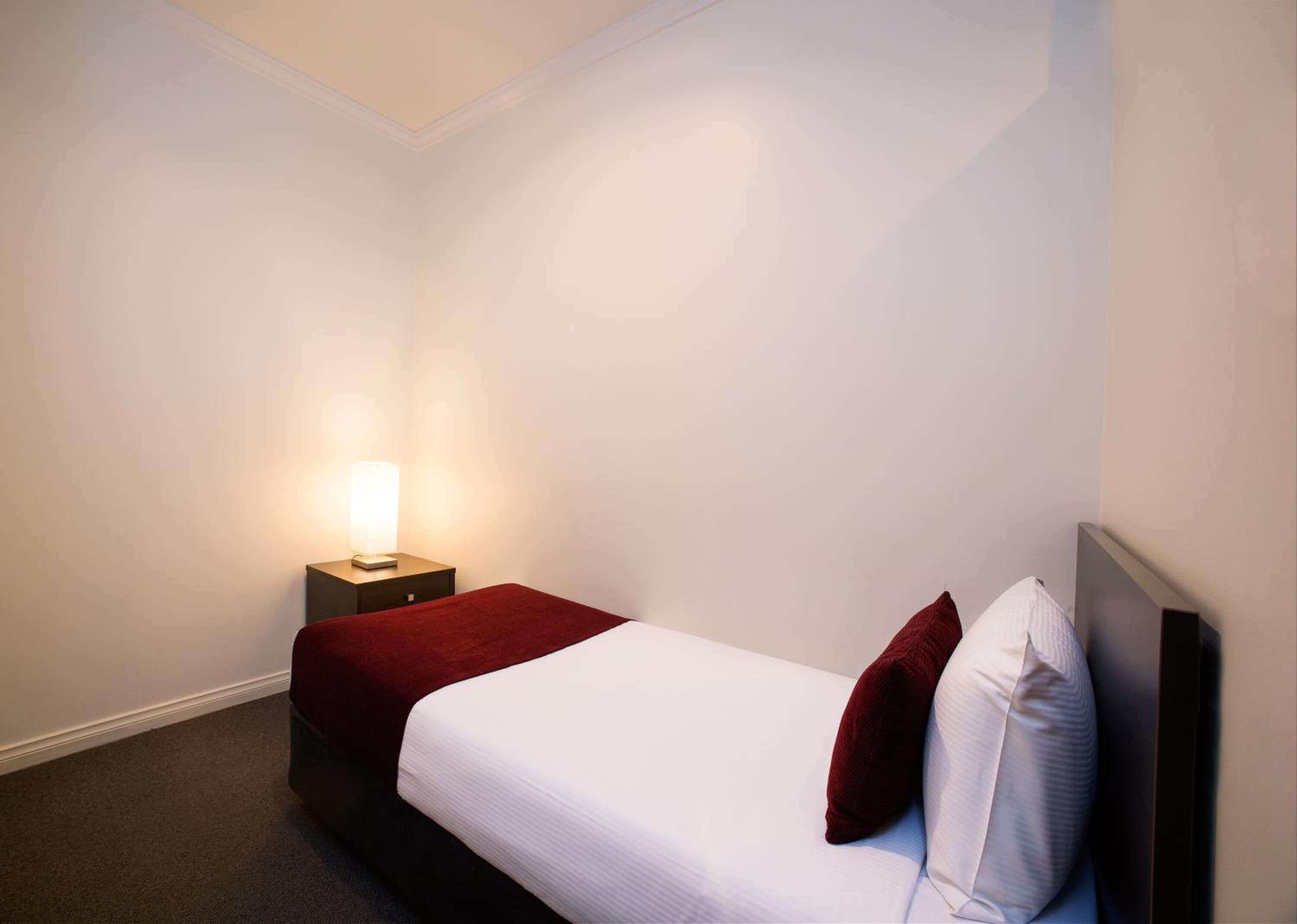 A hotel room with a single bed, a bedside table with a lamp, and a red throw blanket.
