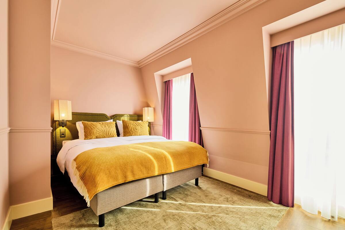 A hotel room with a bed, two bedside lamps, a window with curtains, and a carpeted floor.