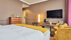 Et hotelværelse med seng, skrivebord med lampe, fjernsyn og et siddeområde med stole.
