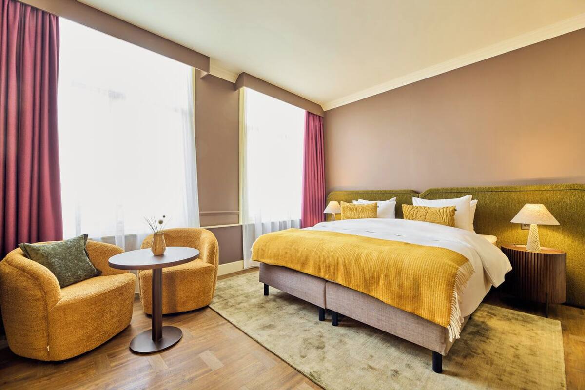 A hotel room with a bed, two armchairs, a small table, and a nightstand with a lamp.