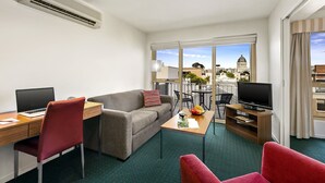 Una habitación de hotel moderna con escritorio, laptop, sillas rojas, sofá gris, televisor y balcón con vistas a la ciudad.