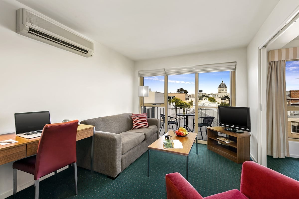 Een moderne hotelkamer met een bureau, laptop, rode stoelen, een grijze bank, een televisie en een balkon met uitzicht op de stad.