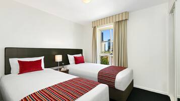 Quarto de hotel com duas camas, cada uma com colcha listrada, travesseiro vermelho e mesa de cabeceira com abajur.