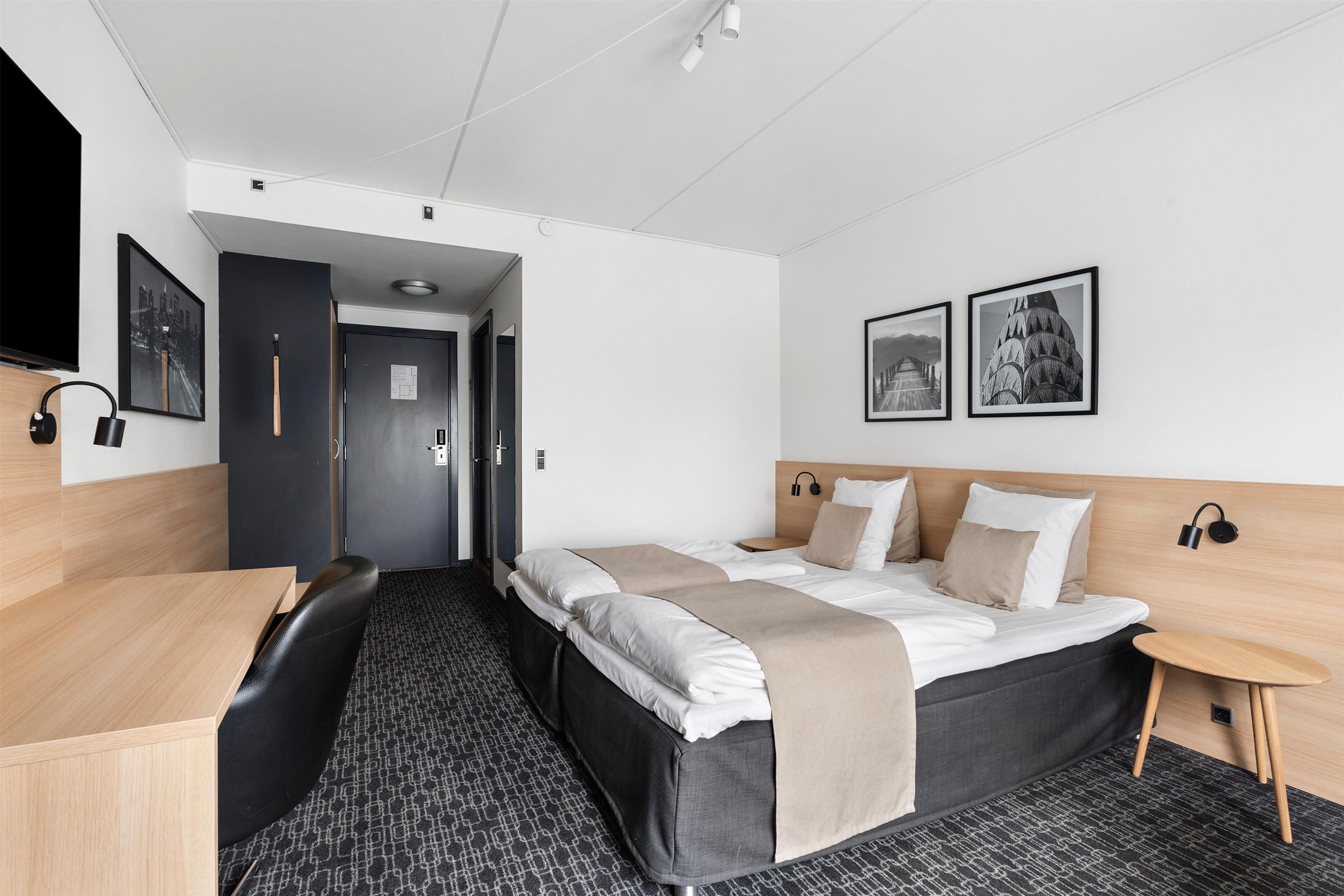 A modern hotel room with a large bed, a desk, and a chair. There are two framed pictures on the wall.