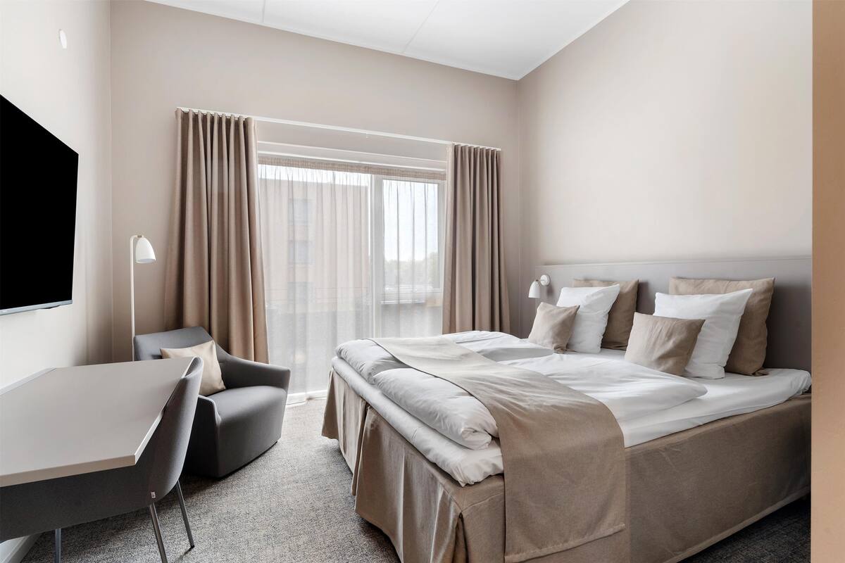 A hotel room with a bed, a desk, a chair, a TV, and a window with curtains.