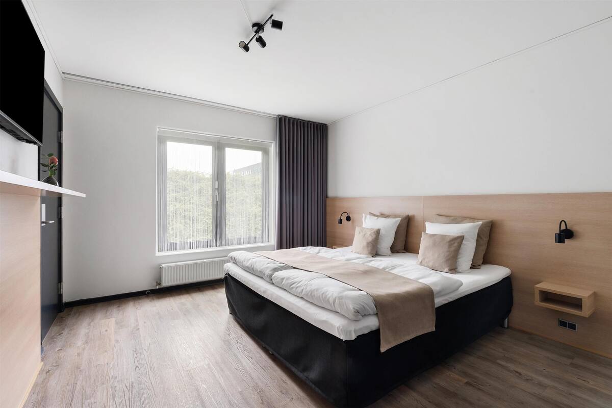 A modern bedroom with a large bed, a window with curtains, a radiator, and a wooden headboard.