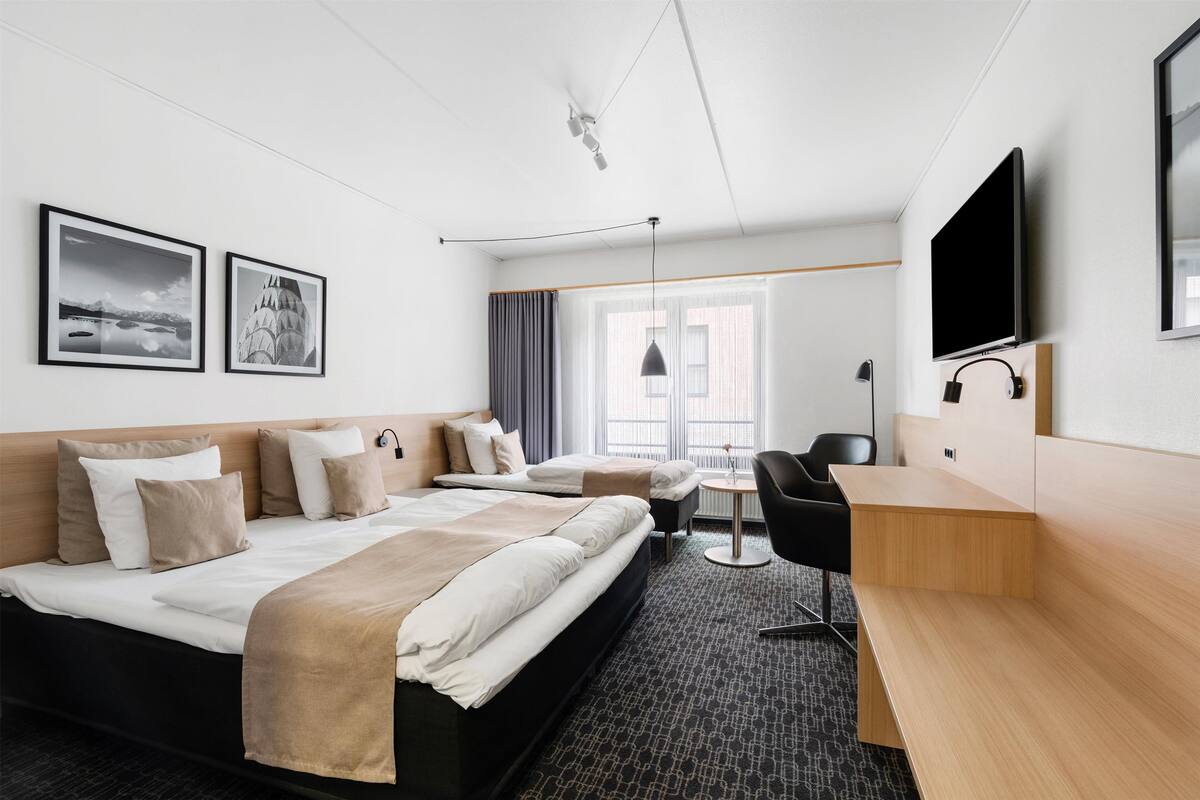 A hotel room with a large bed, a desk, a chair, and a TV. There are two framed pictures on the wall and a window with curtains.