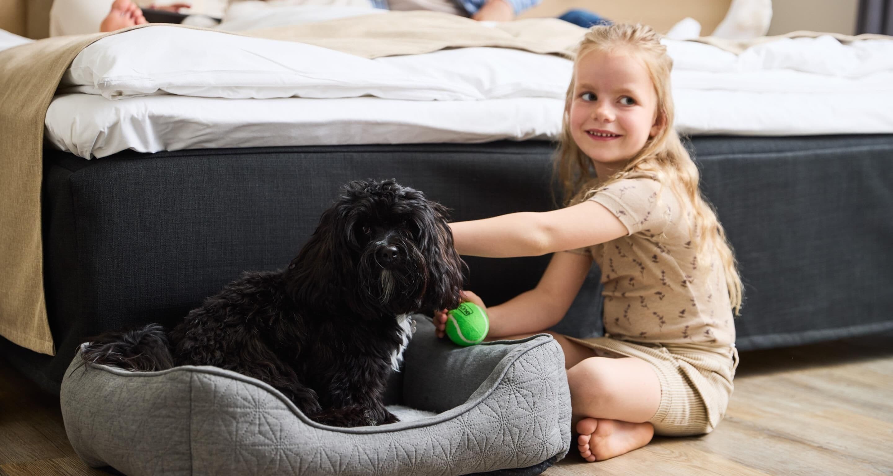 A child sitting on the floor next to a dog in a pet bed, with a bed and a person in the background.