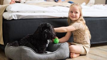 A child sitting on the floor next to a dog in a pet bed, with a bed and a person in the background.