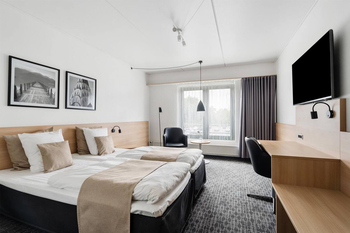 A hotel room with a bed, a desk, a chair, a TV, and two framed pictures on the wall.