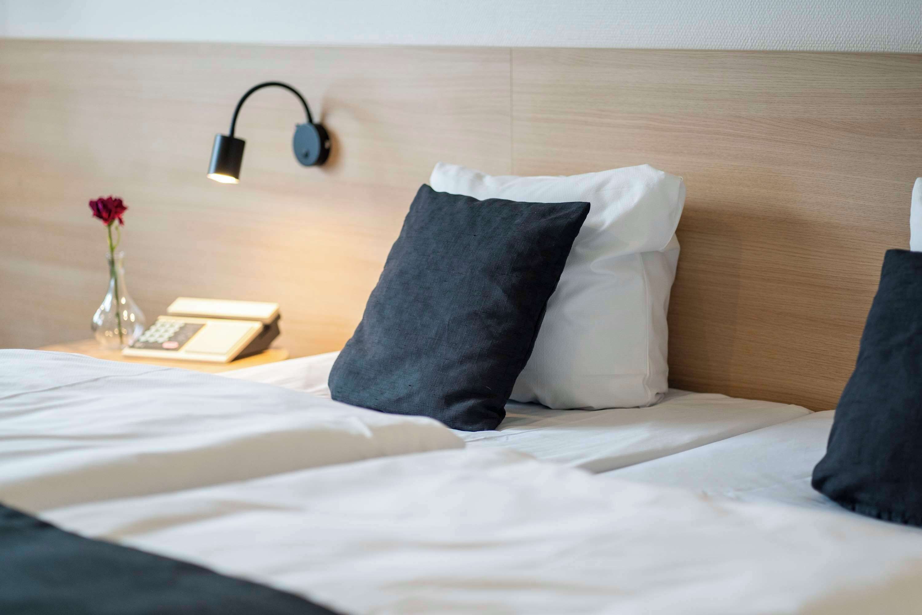 Una cama bien hecha con almohadas blancas y negras, una mesita de noche con una lámpara, un jarrón con una flor roja y un teléfono.