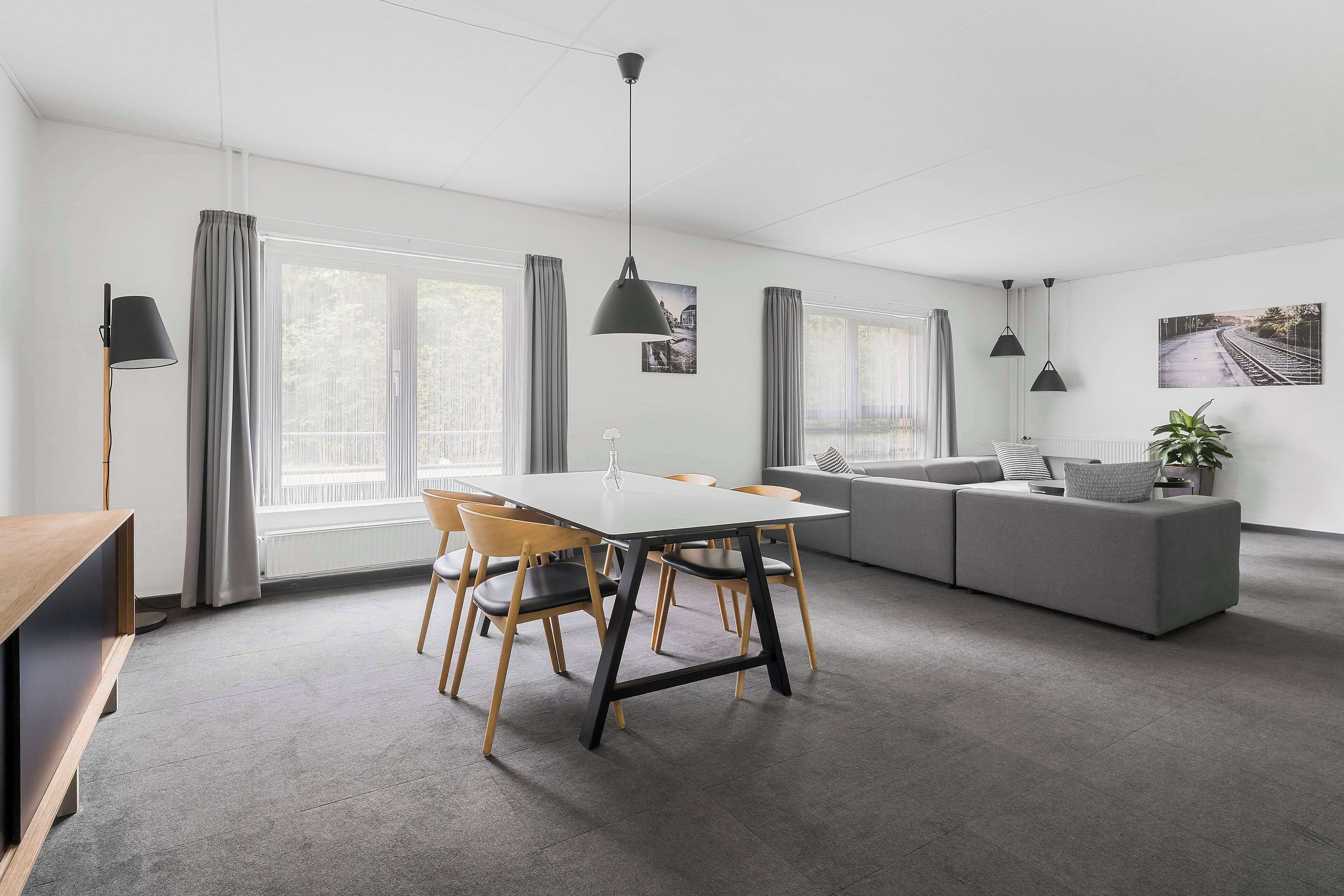 A modern living room with a dining area, grey sofa, and a large window with curtains.
