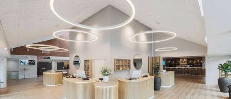 Un área de recepción moderna con mostradores de madera, un bar al fondo y un techo alto con luminarias circulares.