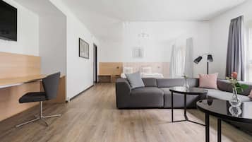 Una habitación de hotel moderna con suelo de madera, un sofá gris, una silla negra, una mesa de centro de cristal y una mesita de noche con lámpara.