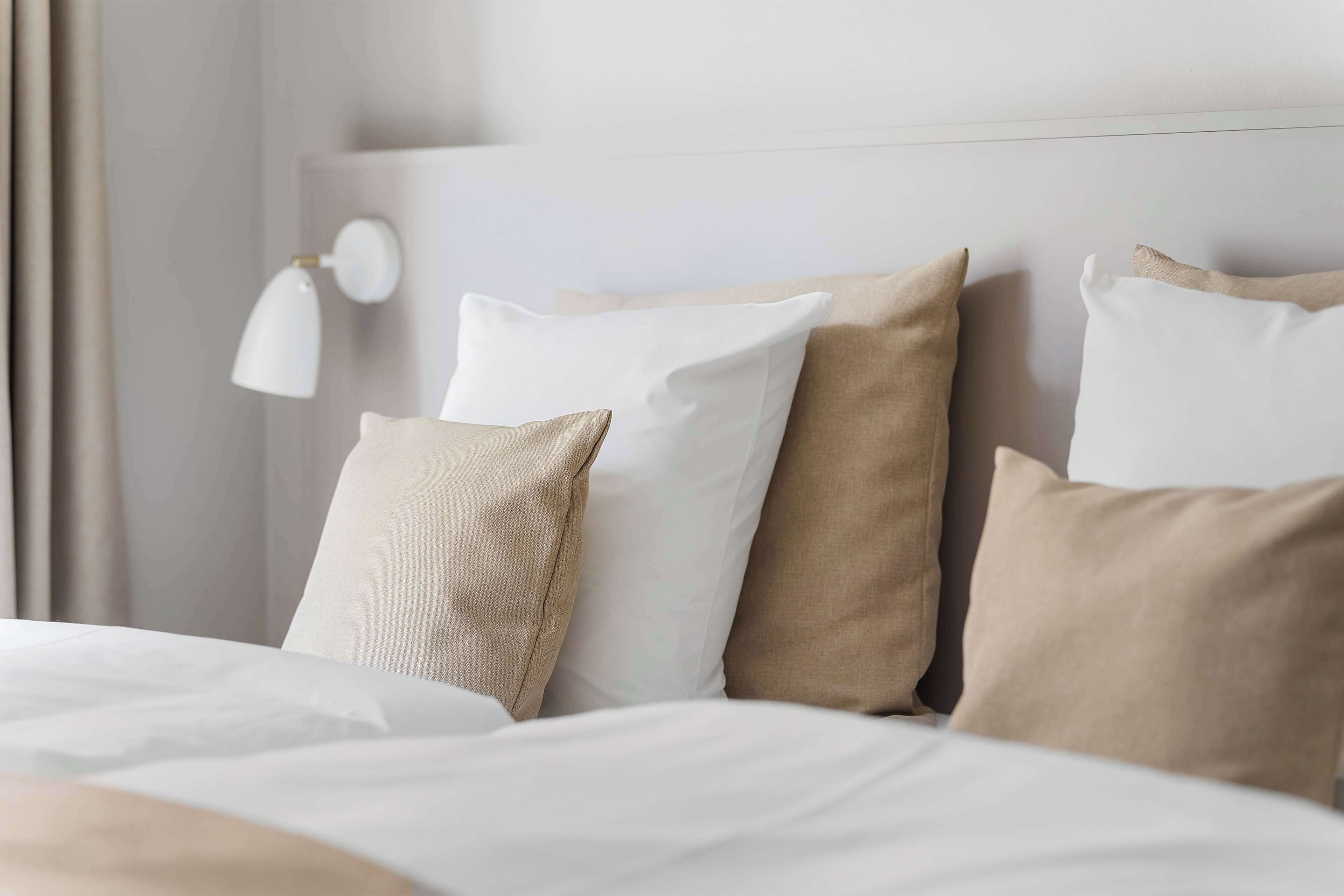 Ein ordentlich bezogenes Bett mit weißer und beiger Bettwäsche, eine Wandlampe und eine schlichte Wand.
