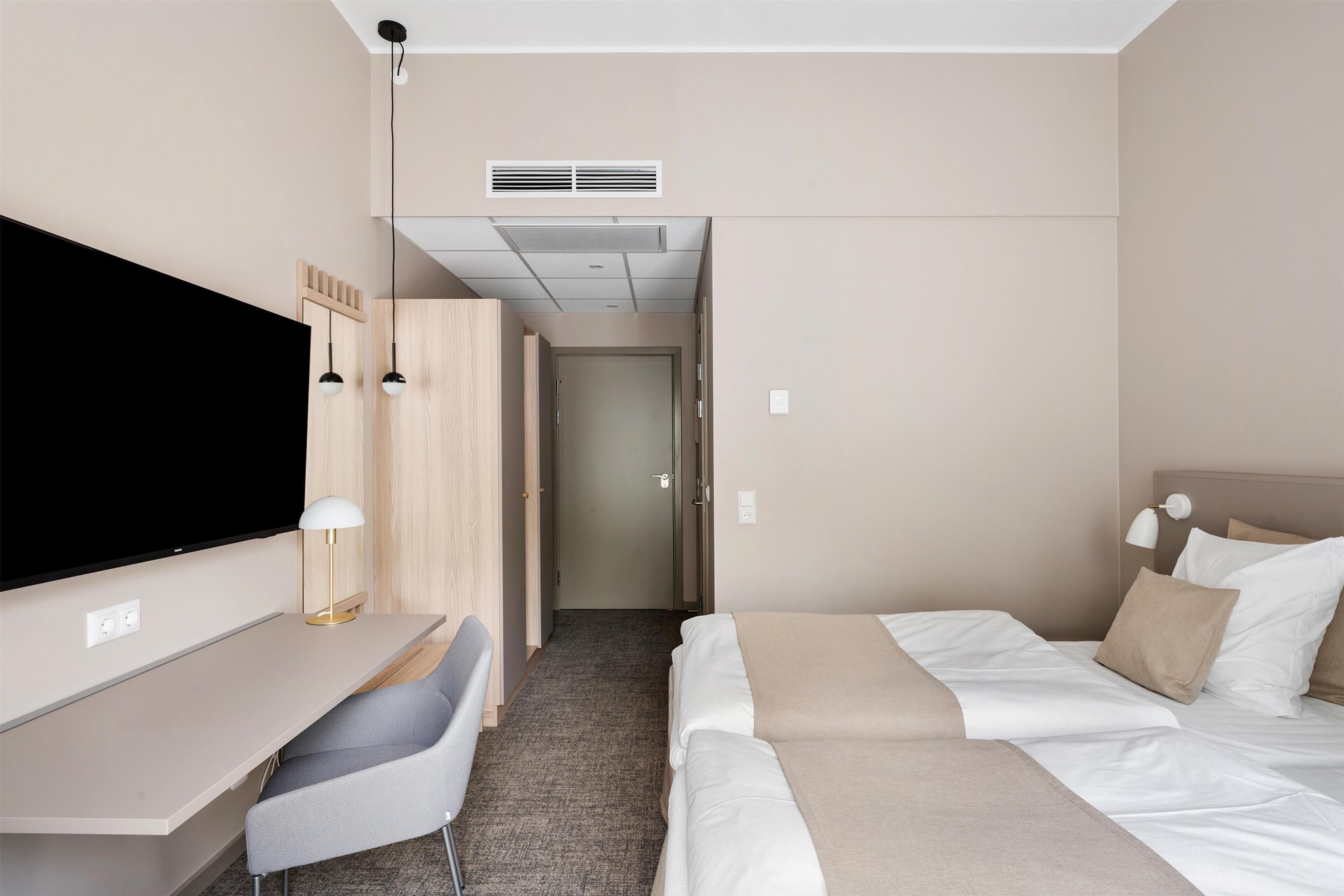 A modern hotel room with a flat-screen TV, a desk with a lamp, a bed with two pillows, and a carpeted floor.