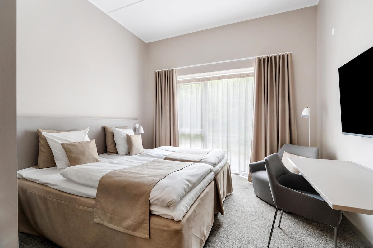 A hotel room with two beds, a desk, and a large window with curtains.