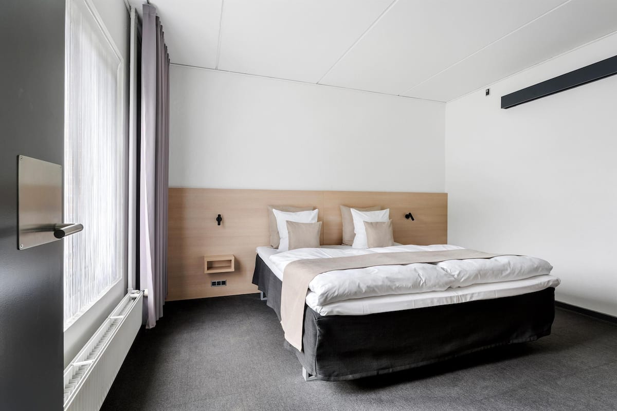 A modern hotel room with a large bed, a bedside table, a built-in shelf, and a window with curtains.